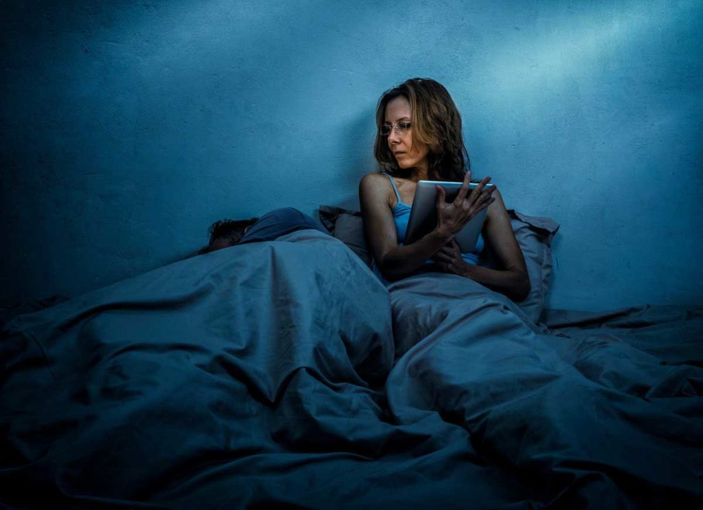 A woman sits up in bed at night using a tablet next to a sleeping man