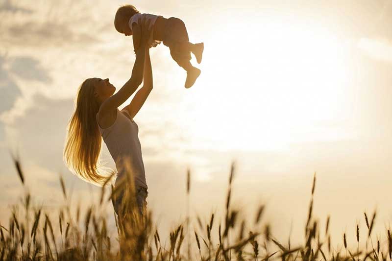 A mother holds her young child in the air in a field, smiling