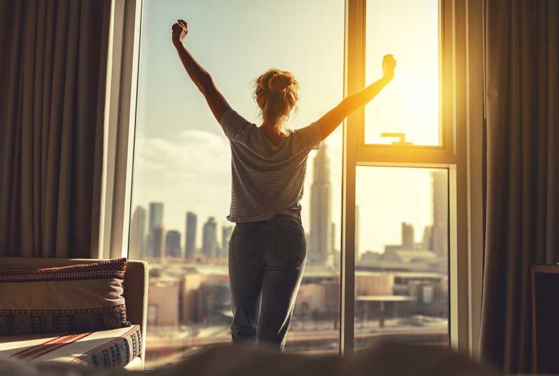 A woman in pajamas stretches in front of a large window while the sun rises